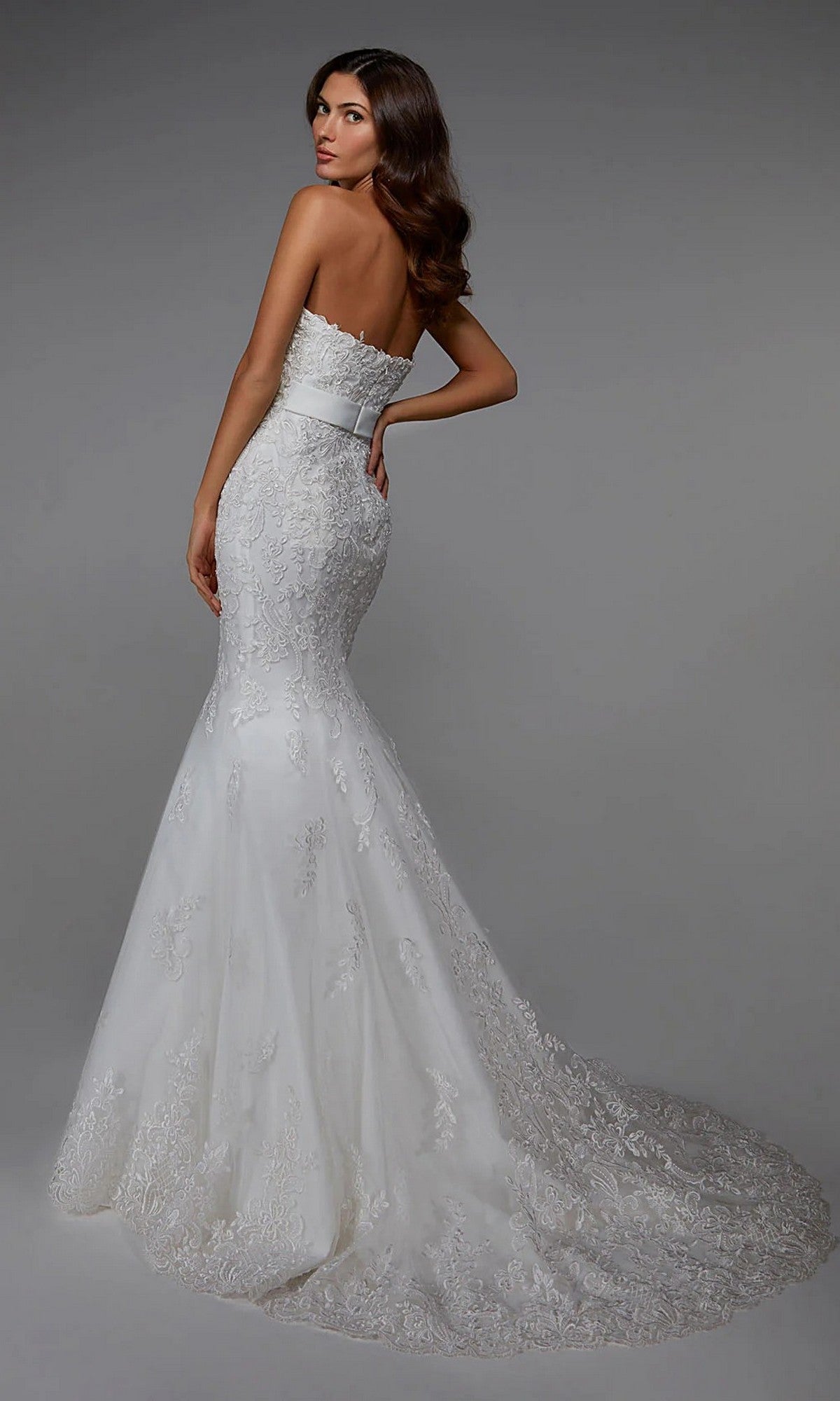 A woman models the Alyce 7032, a long white strapless bridal gown with lace details, a fitted mermaid tulle skirt, and a sweeping train, standing against a plain gray background.